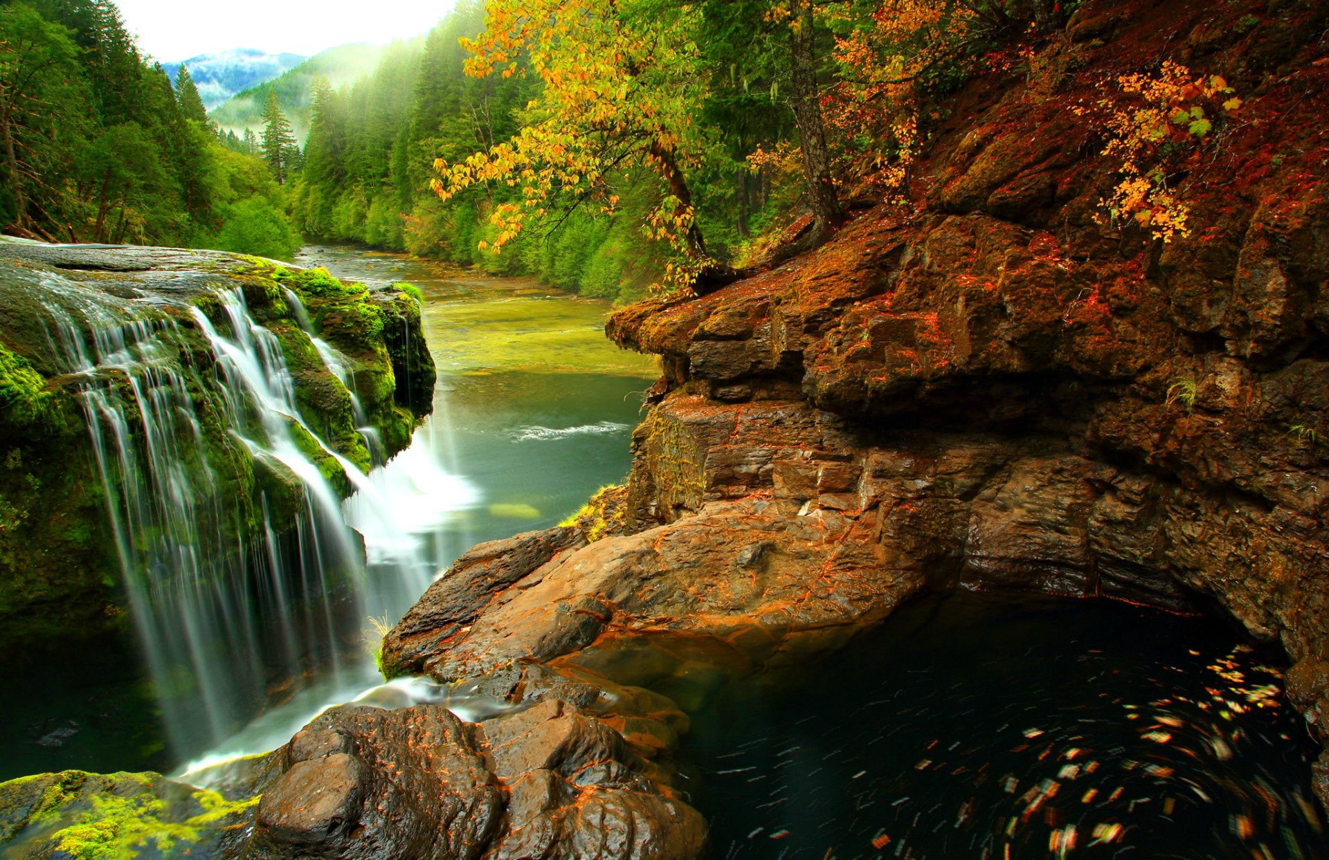 HD desktop wallpaper of a mountain forest with a flowing river and waterfall surrounded by vibrant autumn foliage and rocky terrain in a lush natural landscape.