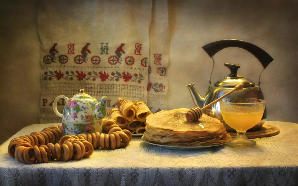 HD still life desktop wallpaper featuring crêpes with honey, a floral teapot, and stacked bagels on a textured table, illuminated by warm, soft lighting.