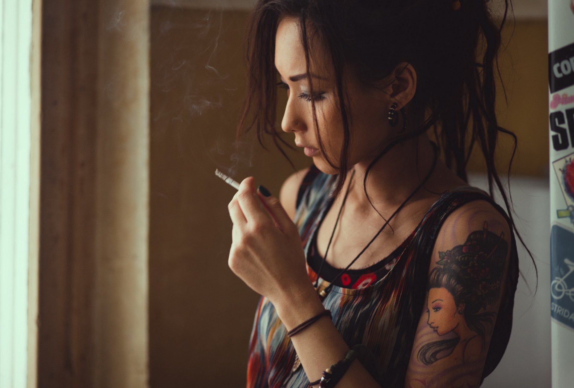 HD wallpaper of a tattooed brunette model smoking. The image captures her in a reflective mood against a soft-lit background.
