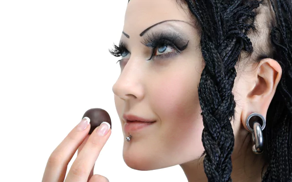 4K Ultra HD PC desktop wallpaper: close-up of a brunette model woman with braided hair, blue eyes, face piercings and a chocolate held near her lips.