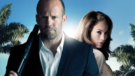 HD desktop wallpaper from the movie Parker showing two actors: a stern bald man in a suit holding a firearm and a woman leaning behind him against palm trees and blue sky.