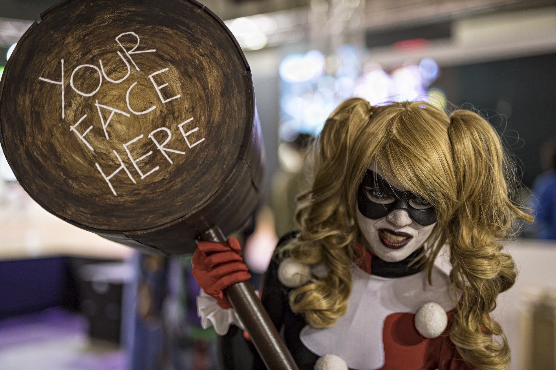 A woman cosplaying Harley Quinn with twintails, blonde hair, blue eyes, and lipstick holds a large mallet with Your Face Here written on it. This image is an HD desktop wallpaper and background.