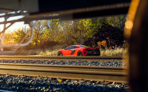 HD desktop wallpaper featuring a vibrant McLaren 675LT supercar parked near railroad tracks, framed by industrial elements and autumn foliage in the background.