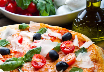 HD desktop wallpaper showing a close-up of a pizza topped with black olives, cherry tomatoes, fresh basil, and shaved cheese, surrounded by ingredients and olive oil.