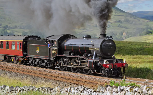 HD wallpaper and background featuring a locomotive pulling a train through scenic countryside. The black train emits smoke as it travels along the track with green hills in the background.