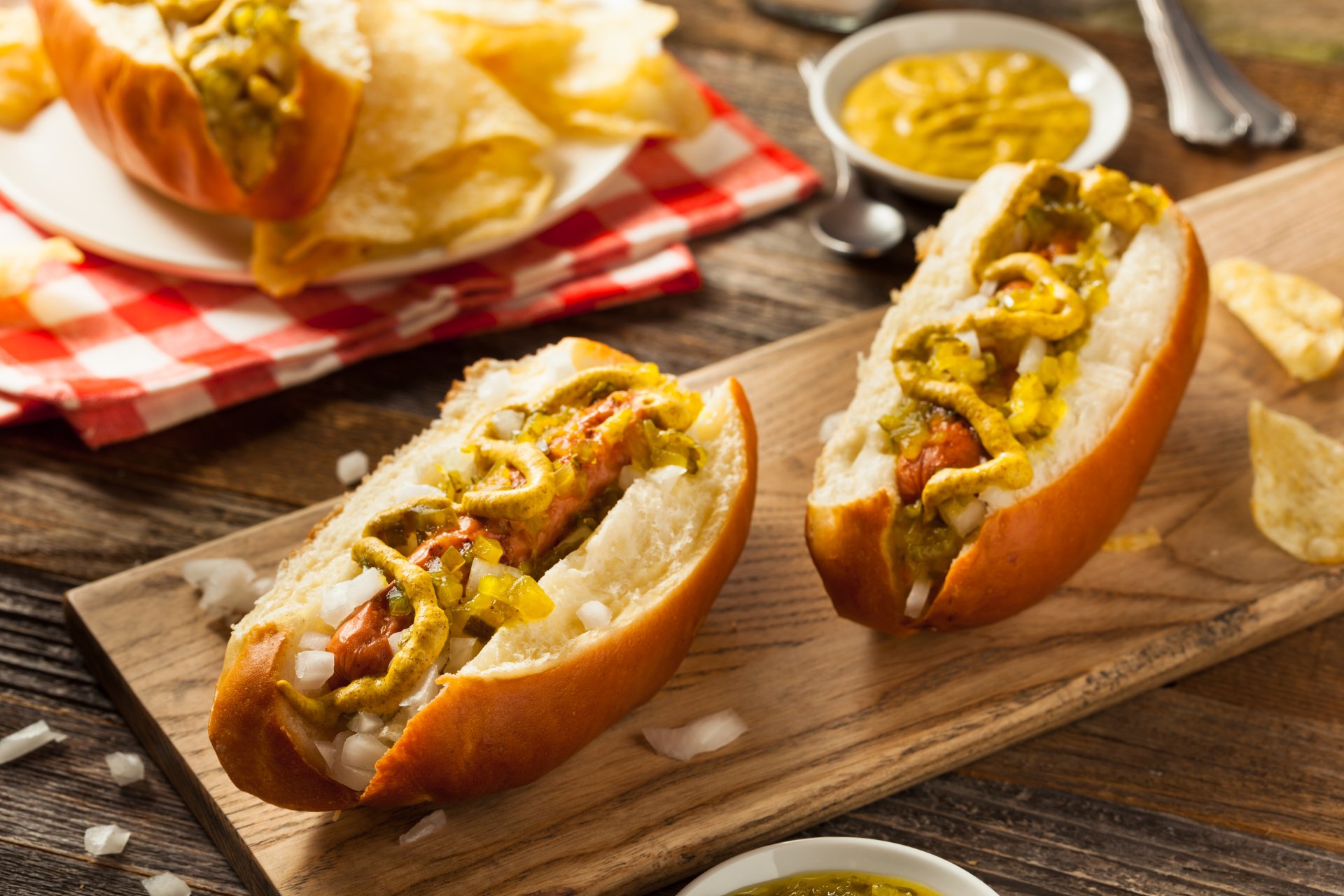 4K Ultra HD PC desktop wallpaper: close-up of two loaded hot dogs on a wooden board with mustard, pickles and potato chips on a rustic table.
