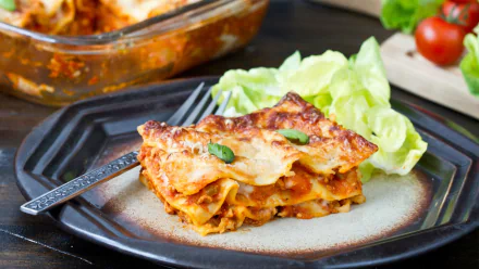 HD desktop wallpaper showing a plated serving of lasagna with melted cheese, accompanied by fresh lettuce on a dark patterned plate.