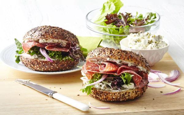 Close-up of two hearty bagel sandwiches with fresh greens and deli meat, accompanied by a bowl of creamy spread and a small salad, captured as an HD PC desktop wallpaper.