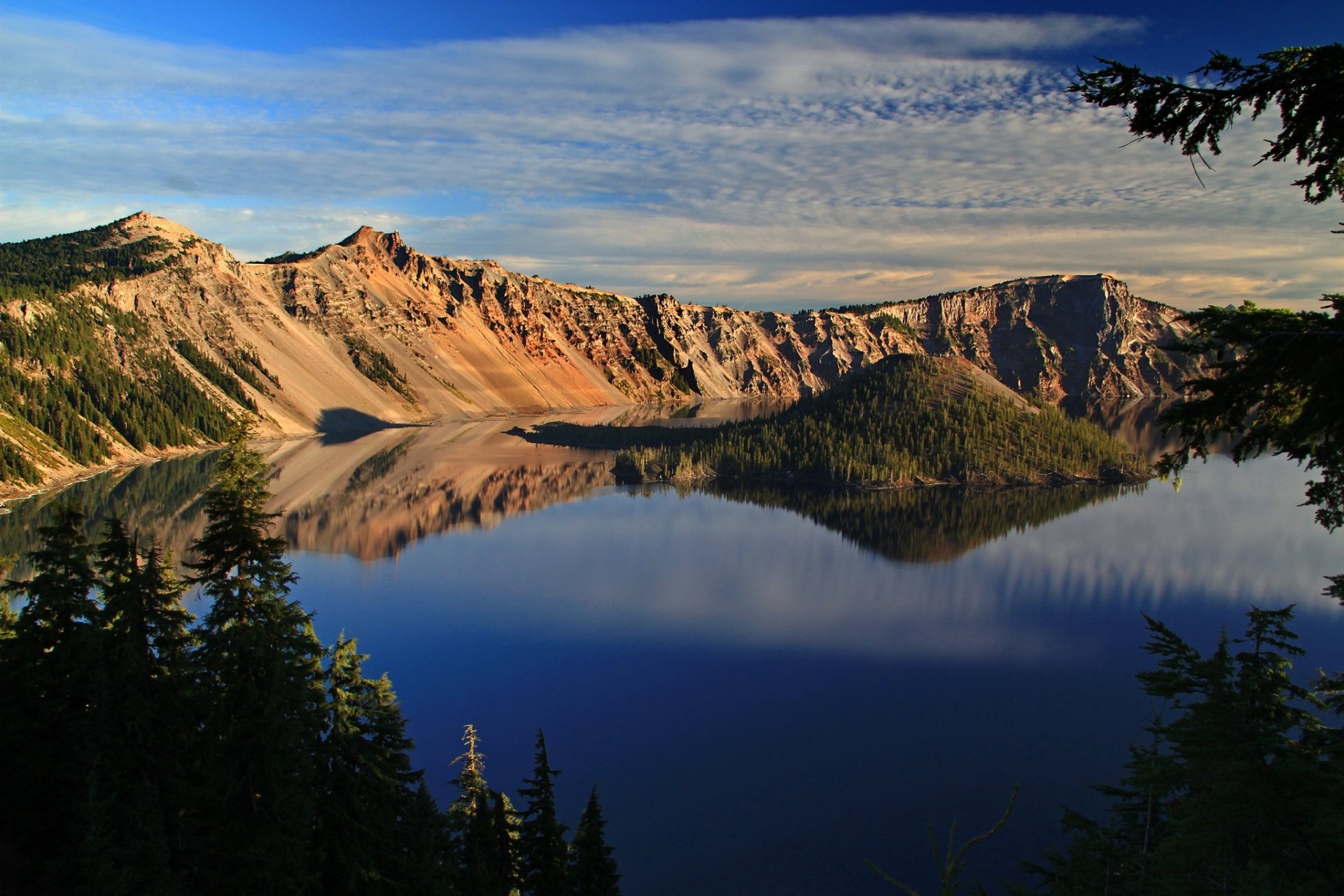 Crystal-clear reflection of forested island and rugged cliffs in the serene waters of Crater Lake, captured in a high-definition nature scene.