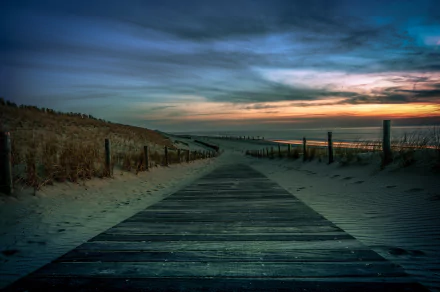 HD desktop wallpaper showcasing a wooden boardwalk leading through sandy dunes toward the ocean horizon under a dramatic sunset sky.