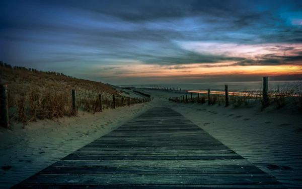 HD desktop wallpaper showcasing a wooden boardwalk leading through sandy dunes toward the ocean horizon under a dramatic sunset sky.