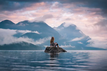 A woman sits on a rock in a calm lake, surrounded by misty mountains under a soft, moody sky, captured in an HD landscape desktop wallpaper.