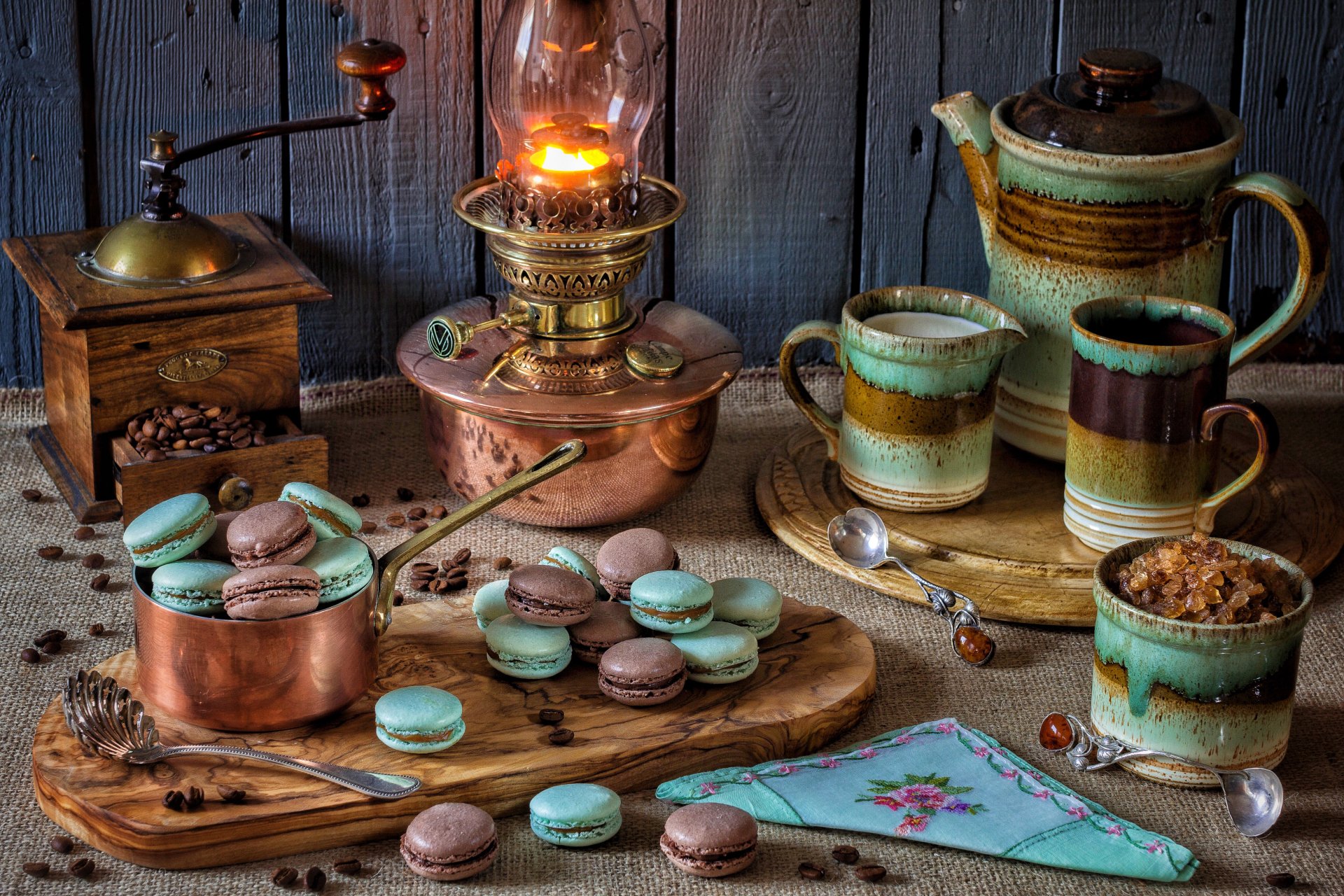 A 4K Ultra HD still life PC wallpaper featuring a vintage coffee grinder, macarons, rustic ceramic cups, and a glowing oil lamp on wooden surfaces.