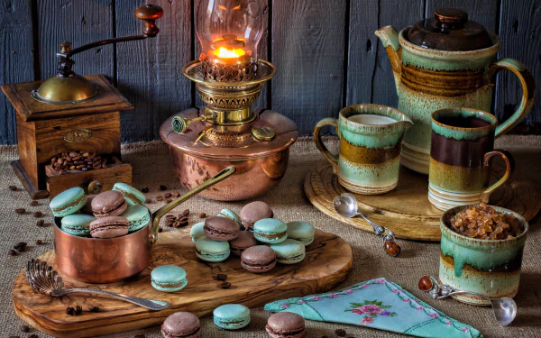 A 4K Ultra HD still life PC wallpaper featuring a vintage coffee grinder, macarons, rustic ceramic cups, and a glowing oil lamp on wooden surfaces.