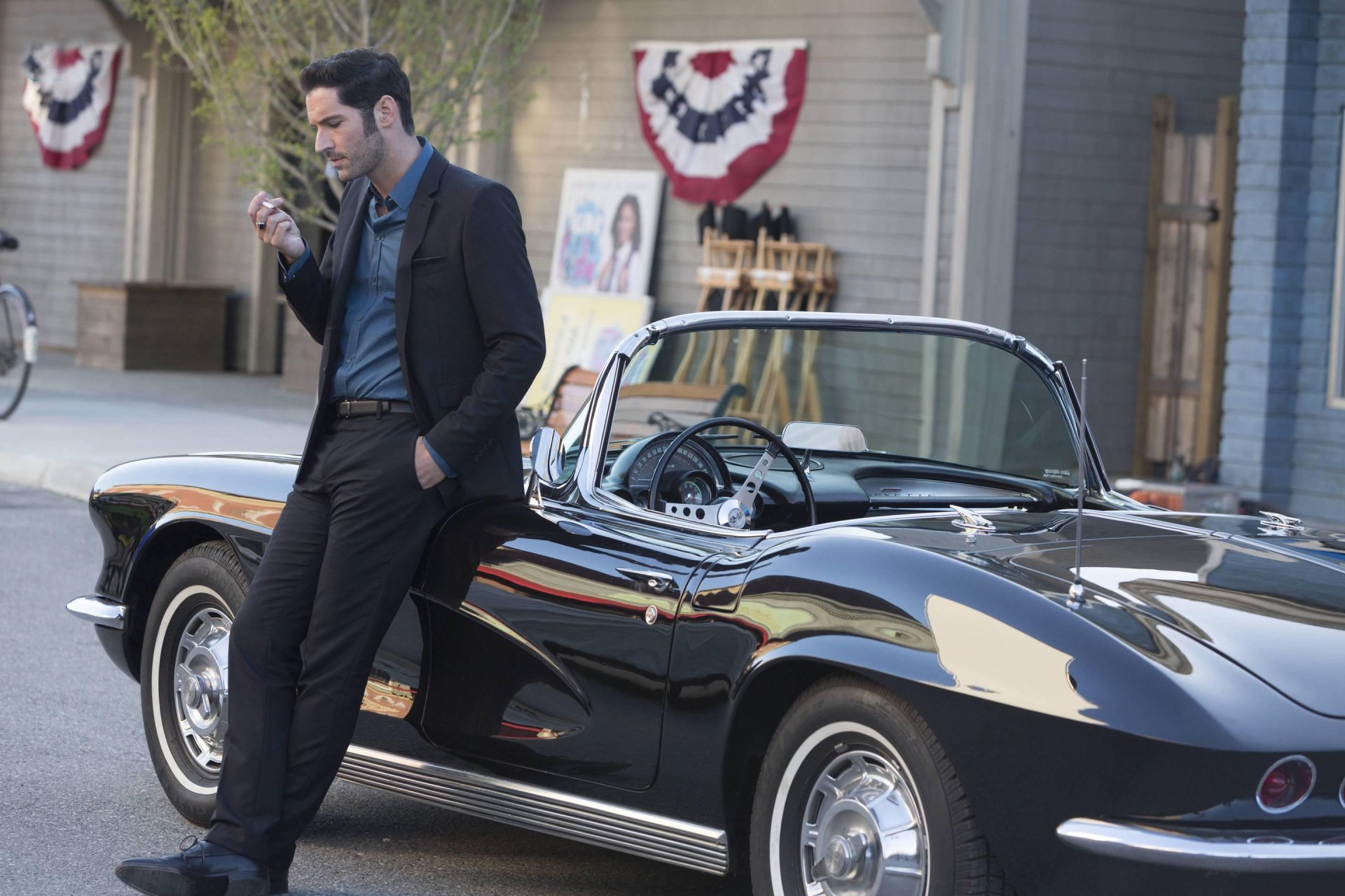 HD wallpaper from the TV show Lucifer featuring Tom Ellis as Lucifer Morningstar, leaning on a classic black car and holding a phone.