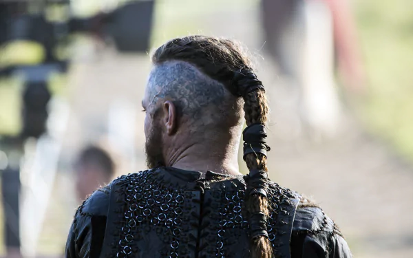 Vikings (2013) TV show 2K Quad HD PC wallpaper — rear view of a braided, partially shaved Viking warrior in chainmail and leather, standing outdoors with blurred figures in the background.
