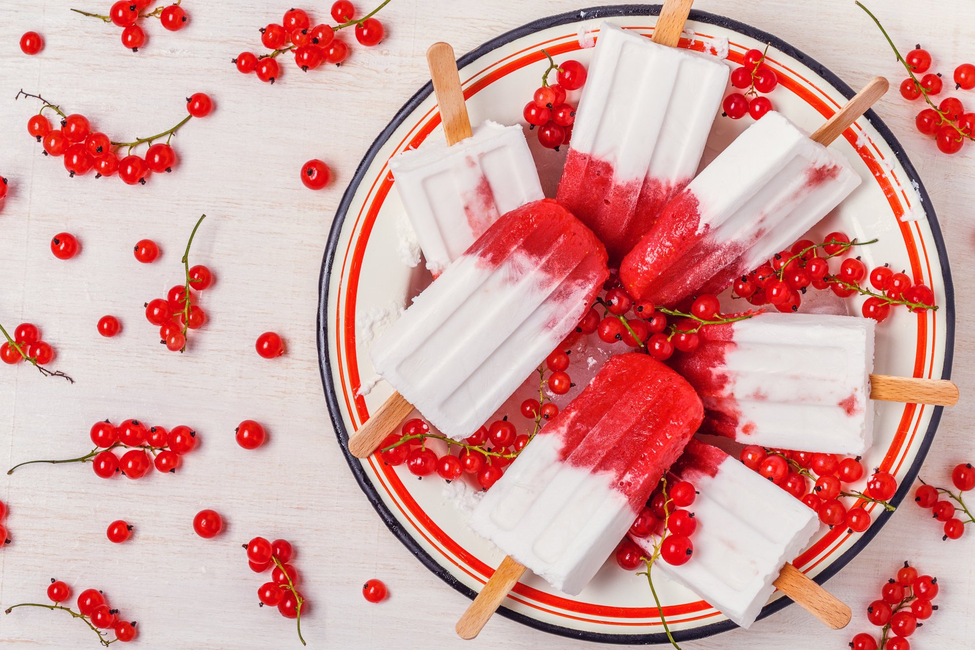 A 4K Ultra HD still life of red currants and creamy white and red ice cream popsicles artfully arranged on a plate and scattered on a white surface.