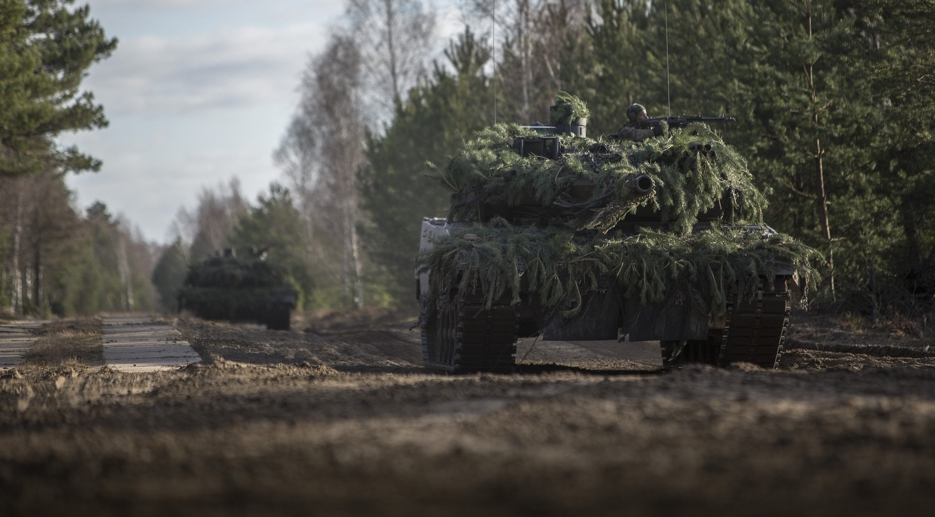 HD desktop wallpaper featuring two Leopard 2 military tanks camouflaged with greenery, moving along a dirt road in a forested area.