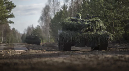 HD desktop wallpaper featuring two Leopard 2 military tanks camouflaged with greenery, moving along a dirt road in a forested area.