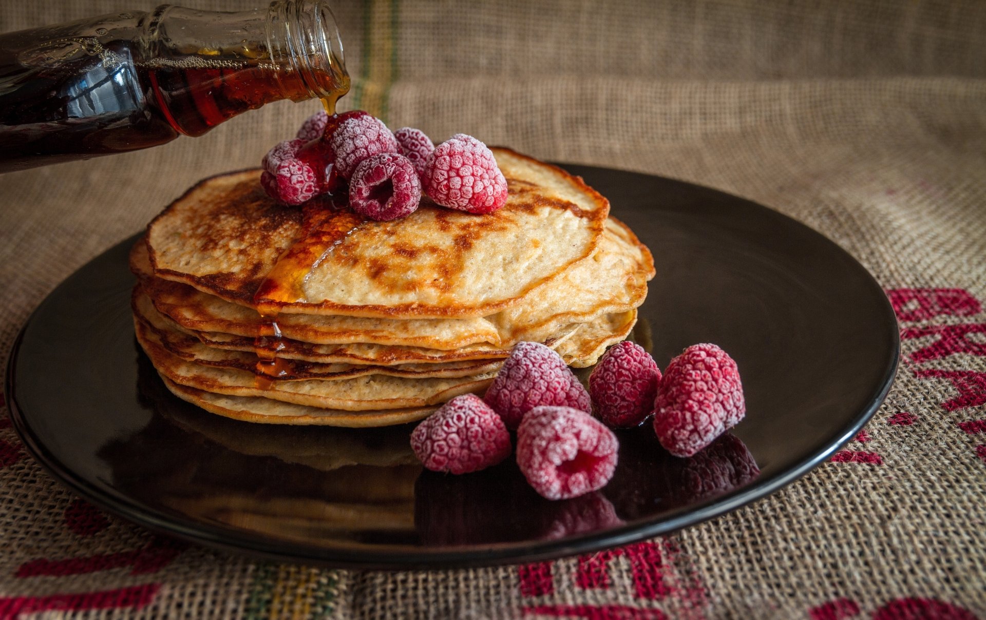 5K Ultra HD PC desktop wallpaper: syrup drizzling over stacked pancakes topped with frozen raspberries, extra berries on a black plate — breakfast food berry close-up