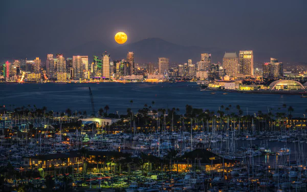 Night view of San Diego harbor with illuminated city skyline, boats in the water, and a glowing full moon rising above the buildings in this 4K Ultra HD desktop wallpaper.