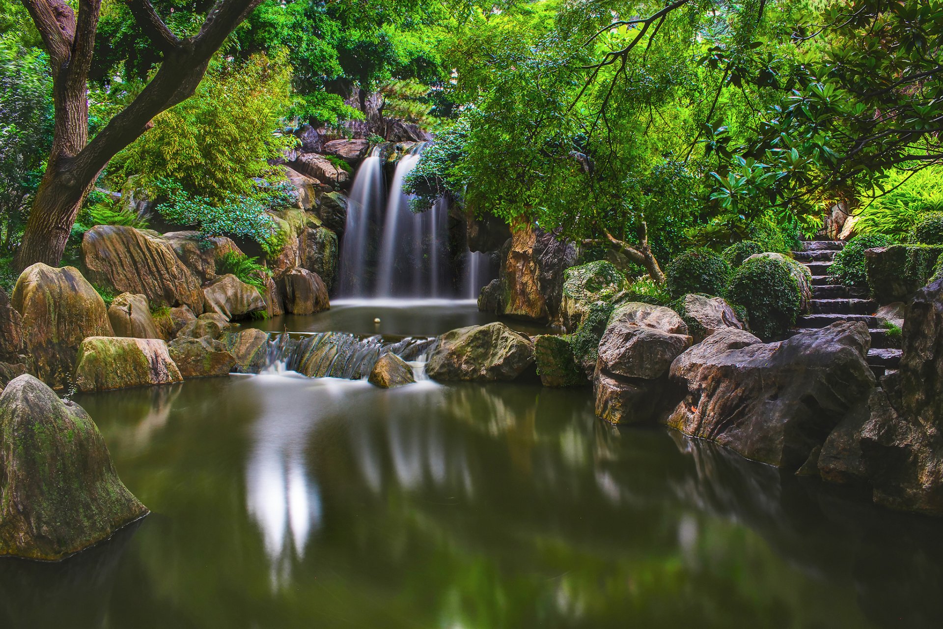 HD desktop wallpaper showing a serene park with a waterfall cascading into a calm pond, framed by lush greenery. Stone stairs are visible among the natural surroundings of the forest.
