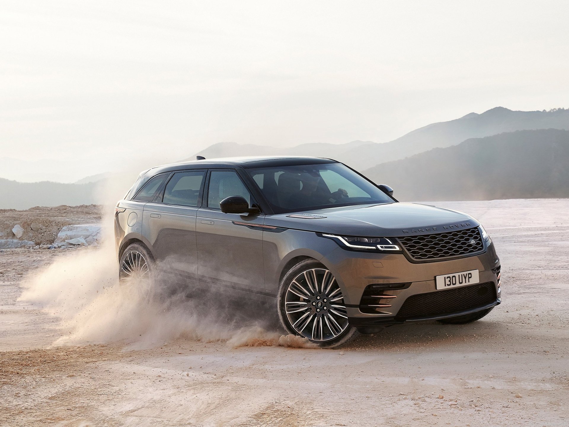 HD desktop wallpaper featuring a silver Range Rover SUV, captured mid-action on a sandy terrain with a mountainous background. The Land Rover vehicle exudes power and elegance.