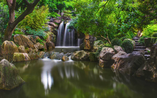HD desktop wallpaper showing a serene park with a waterfall cascading into a calm pond, framed by lush greenery. Stone stairs are visible among the natural surroundings of the forest.