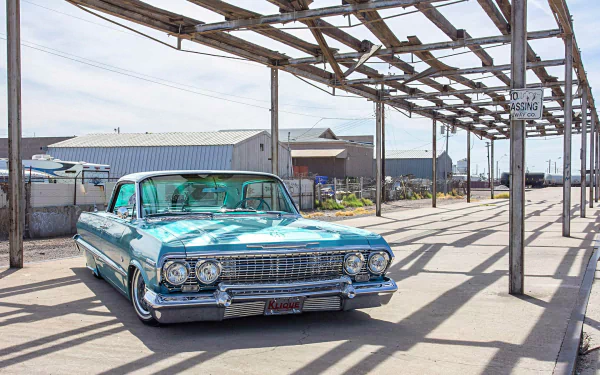 Teal Chevrolet Impala lowrider parked under an open steel canopy at an industrial pier — classic car HD desktop wallpaper background