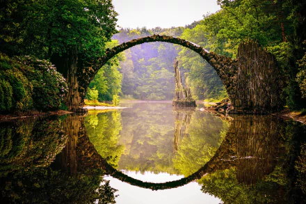 A stunning HD wallpaper of the Devil's Bridge (Rakotzbrücke) in Park Kromlau, Germany, featuring a man-made arch bridge reflecting perfectly in the calm river surrounded by lush green trees.