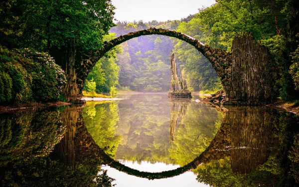 A stunning HD wallpaper of the Devil's Bridge (Rakotzbrücke) in Park Kromlau, Germany, featuring a man-made arch bridge reflecting perfectly in the calm river surrounded by lush green trees.