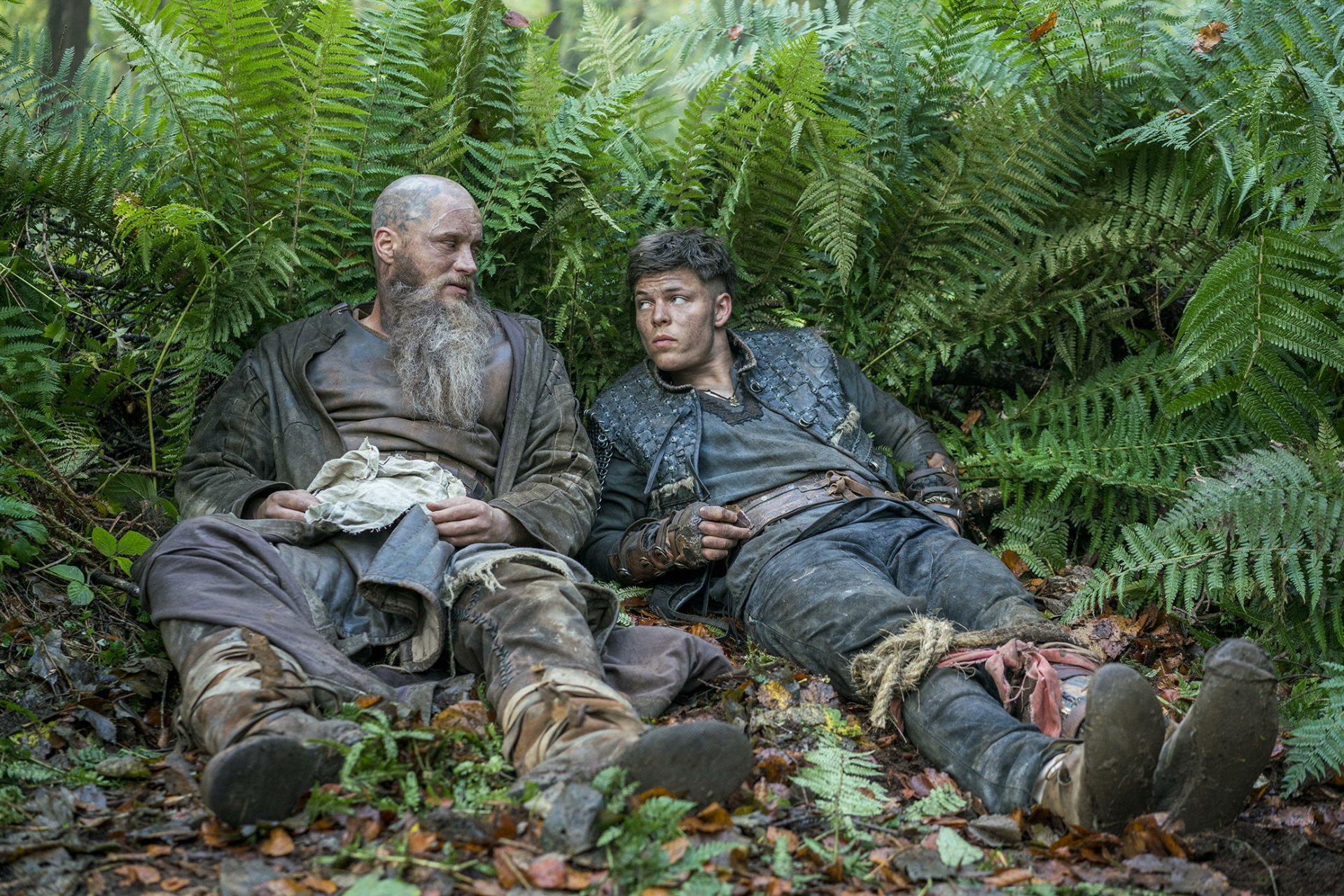 Ivar The Boneless and a bearded man rest against lush ferns in a forest scene from the TV show Vikings (2013), presented as an HD PC desktop wallpaper.