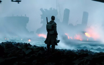 A soldier stands on a rocky shoreline, facing a burning ship in the background, in a high-definition scene from the movie Dunkirk.