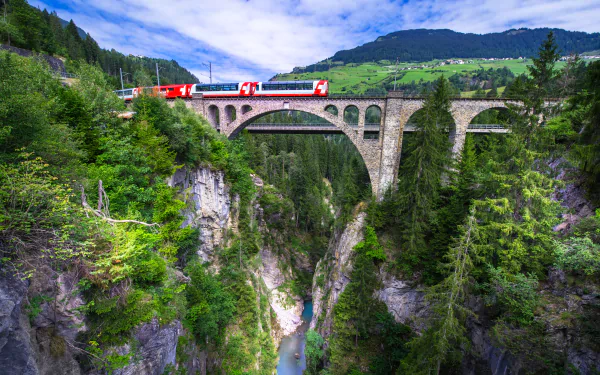 A red train crosses a stone arch bridge over a deep gorge surrounded by lush green forests and hills in Switzerland, captured in 4K Ultra HD.