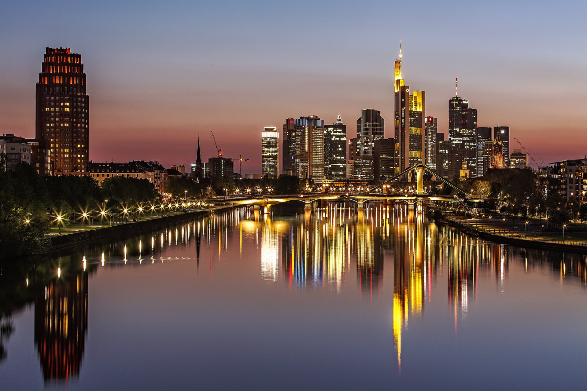 Night view of Frankfurt’s illuminated skyscrapers and city buildings reflecting on the river, featuring bridges and city lights in Germany.