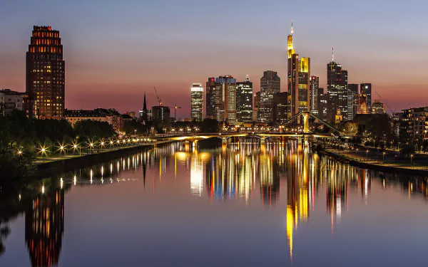 Night view of Frankfurt’s illuminated skyscrapers and city buildings reflecting on the river, featuring bridges and city lights in Germany.