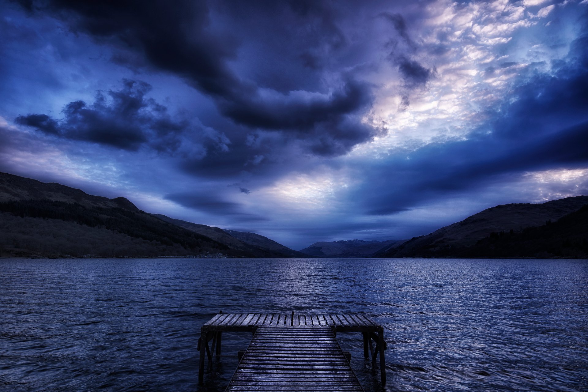 A stunning HD wallpaper featuring a serene lake, a rustic pier extending into the water, and a dramatic sky filled with dark clouds under a blue hue.