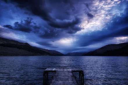 A stunning HD wallpaper featuring a serene lake, a rustic pier extending into the water, and a dramatic sky filled with dark clouds under a blue hue.