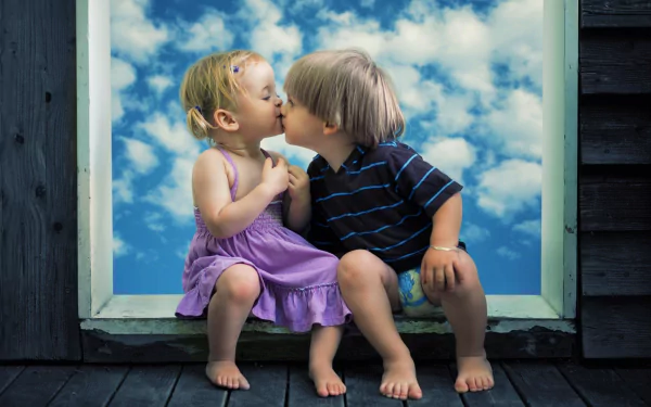 Two children, a little boy and a little girl, share a cute kiss while sitting barefoot against a backdrop of a blue sky filled with fluffy clouds. This HD desktop wallpaper captures a heartwarming moment of childhood.