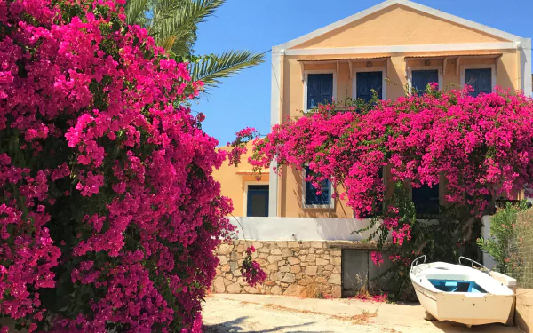 2K Quad HD desktop wallpaper: sunlit Greek stone house draped in vibrant purple bougainvillea, with a small white boat ashore.