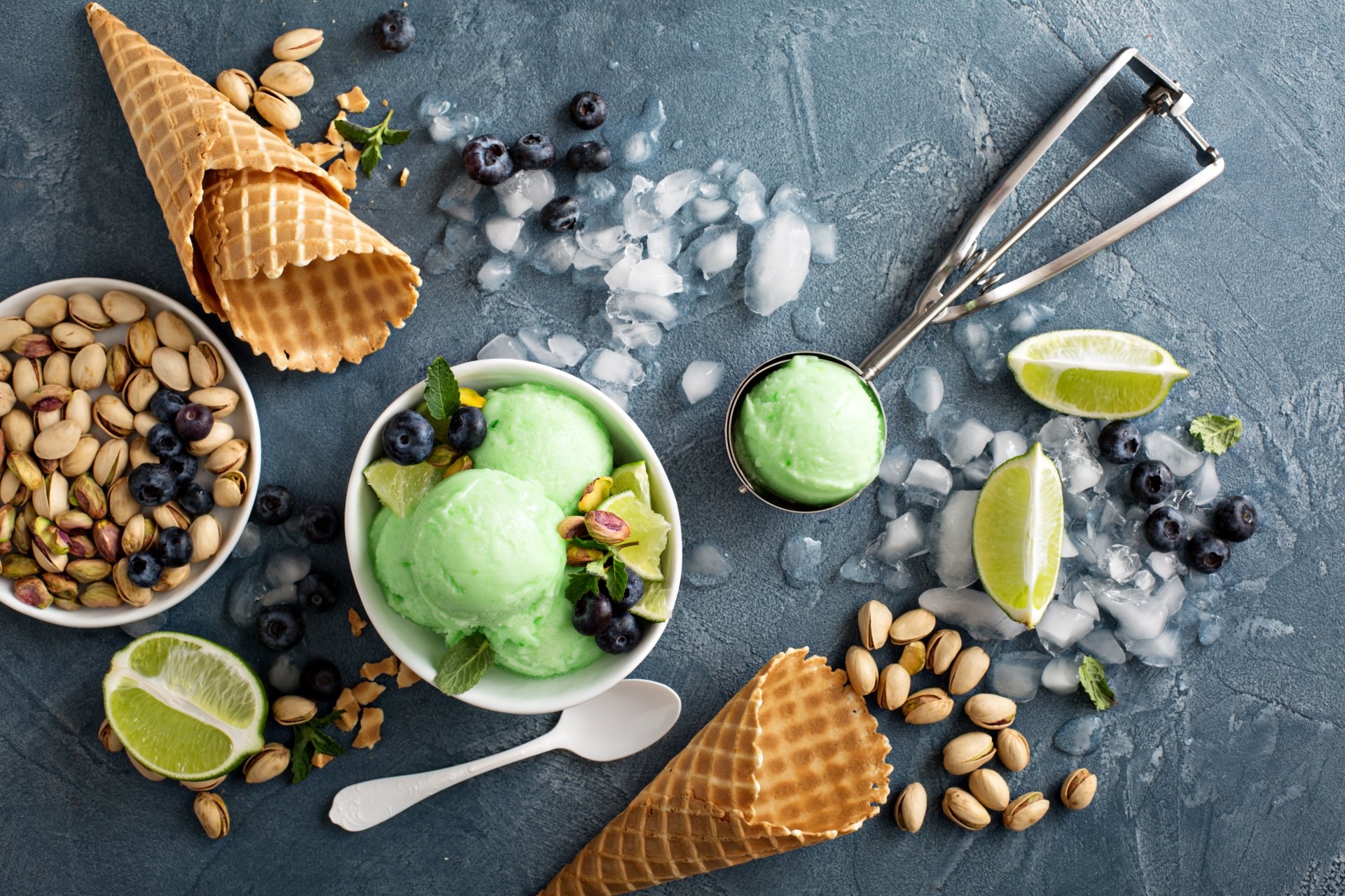 A 4K Ultra HD still life of pistachio ice cream scoops with blueberries, waffle cones, pistachio nuts, lime wedges, and ice cubes on a textured dark surface.