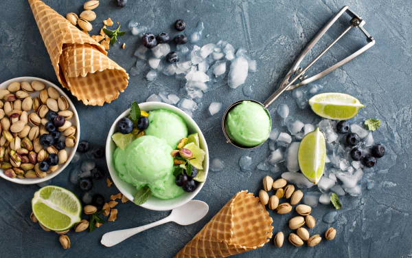 A 4K Ultra HD still life of pistachio ice cream scoops with blueberries, waffle cones, pistachio nuts, lime wedges, and ice cubes on a textured dark surface.