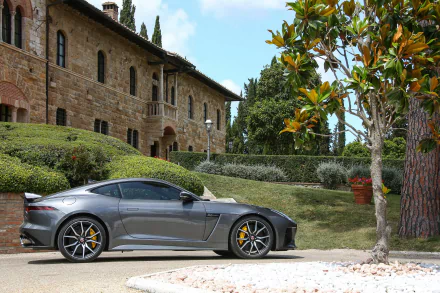 Silver Jaguar F-Type parked beside a garden with manicured bushes and a stone building under a clear sky, captured in 4K Ultra HD quality.