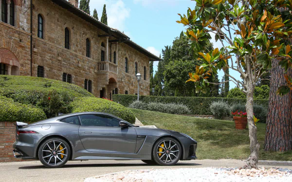 Silver Jaguar F-Type parked beside a garden with manicured bushes and a stone building under a clear sky, captured in 4K Ultra HD quality.