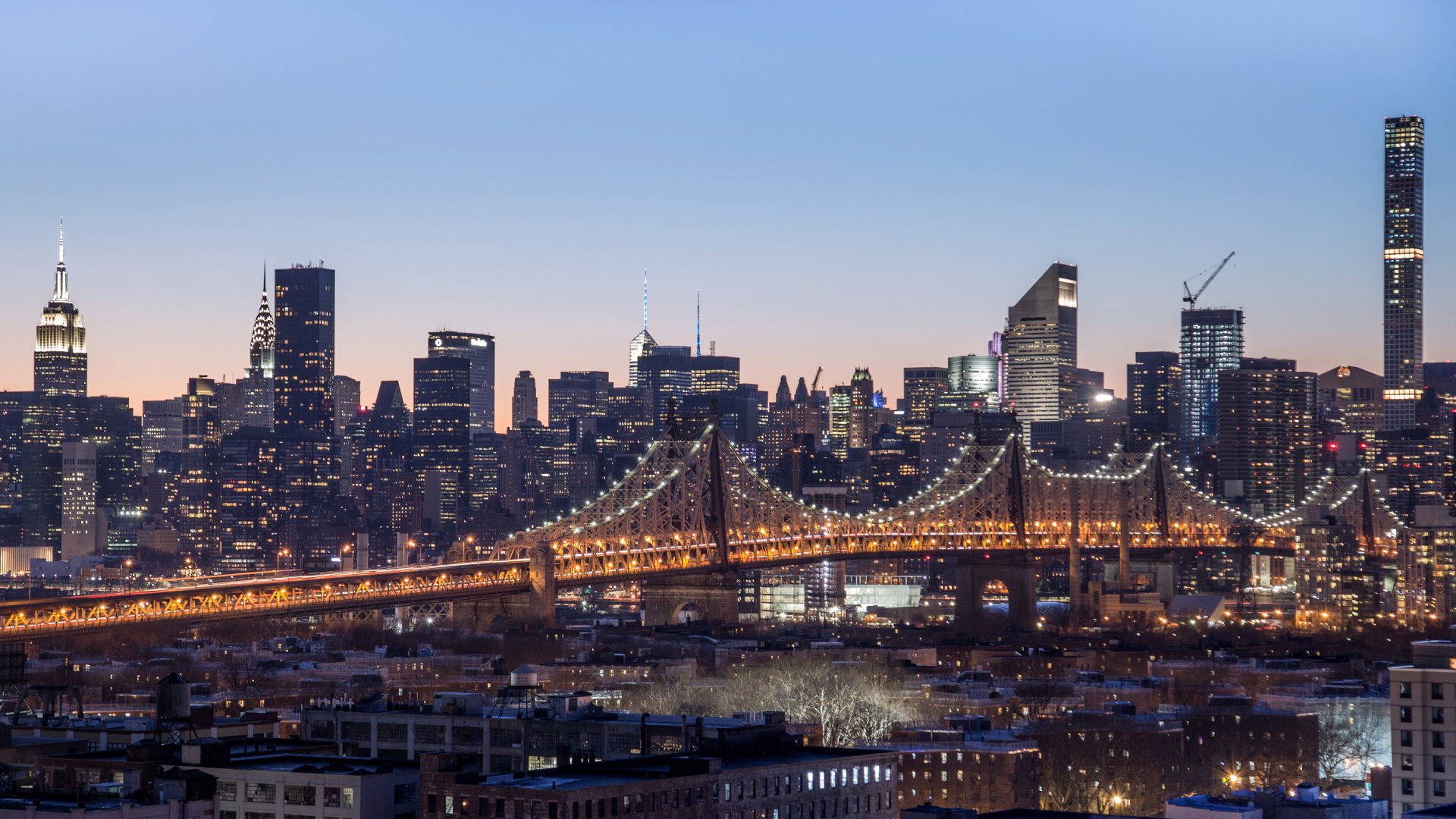 Queensboro Bridge lit at dusk with Manhattan skyscrapers rising behind, New York City skyline, man-made urban panorama, USA; 2K Quad HD PC desktop wallpaper and background.