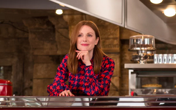 Julianne Moore from Kingsman: The Golden Circle poses behind a diner counter, wearing a red and blue patterned top. This HD desktop wallpaper showcases her character confidently in a classic diner setting.