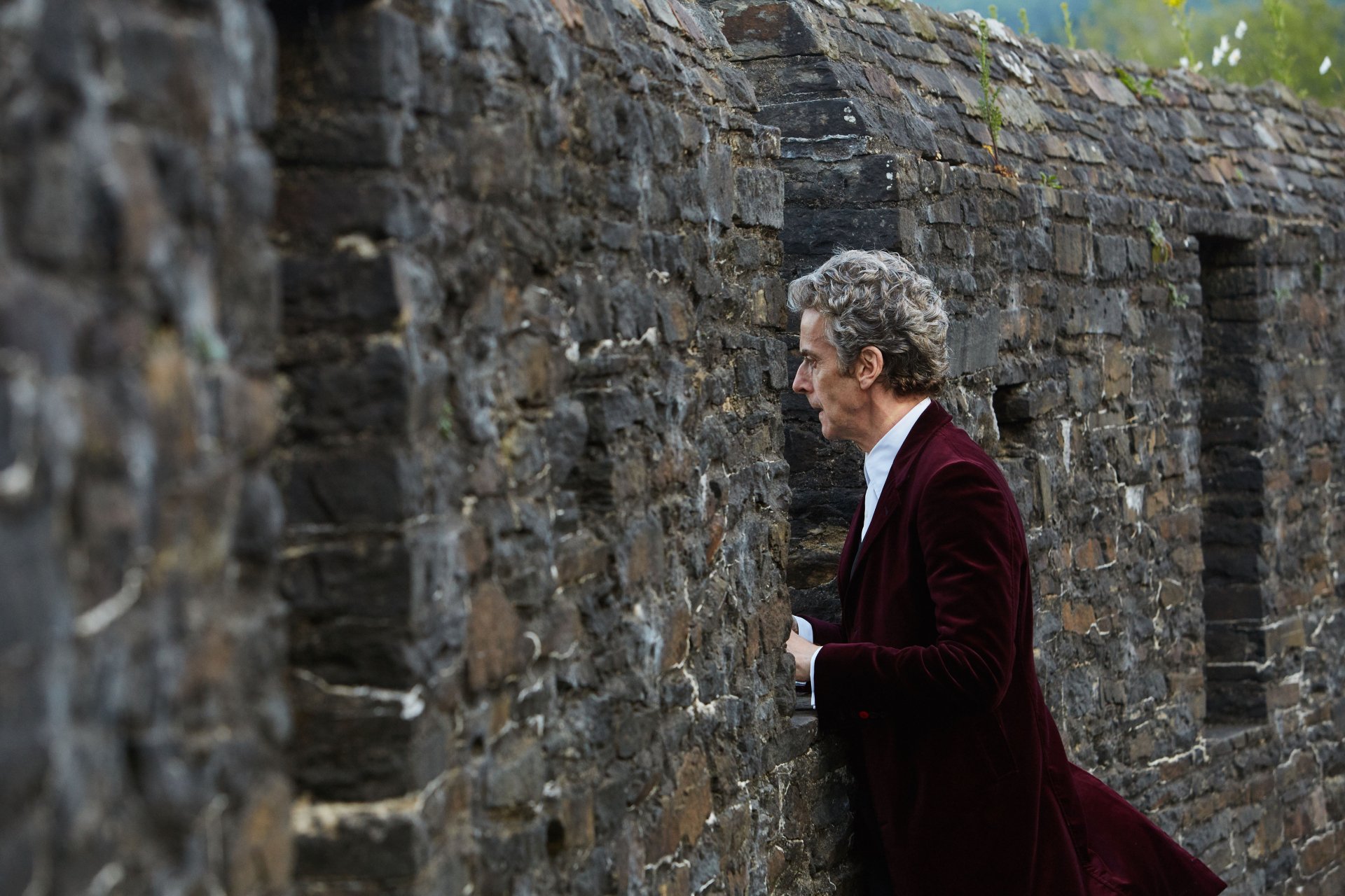 Peter Capaldi as the 12th Doctor in Doctor Who (2005) stands next to a stone wall, captured in this 4K Ultra HD PC desktop wallpaper and background.
