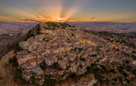 A stunning HD desktop wallpaper of Assoro, Italy, showcasing a man-made village nestled on rugged hills at sunrise, with warm light illuminating the landscape.