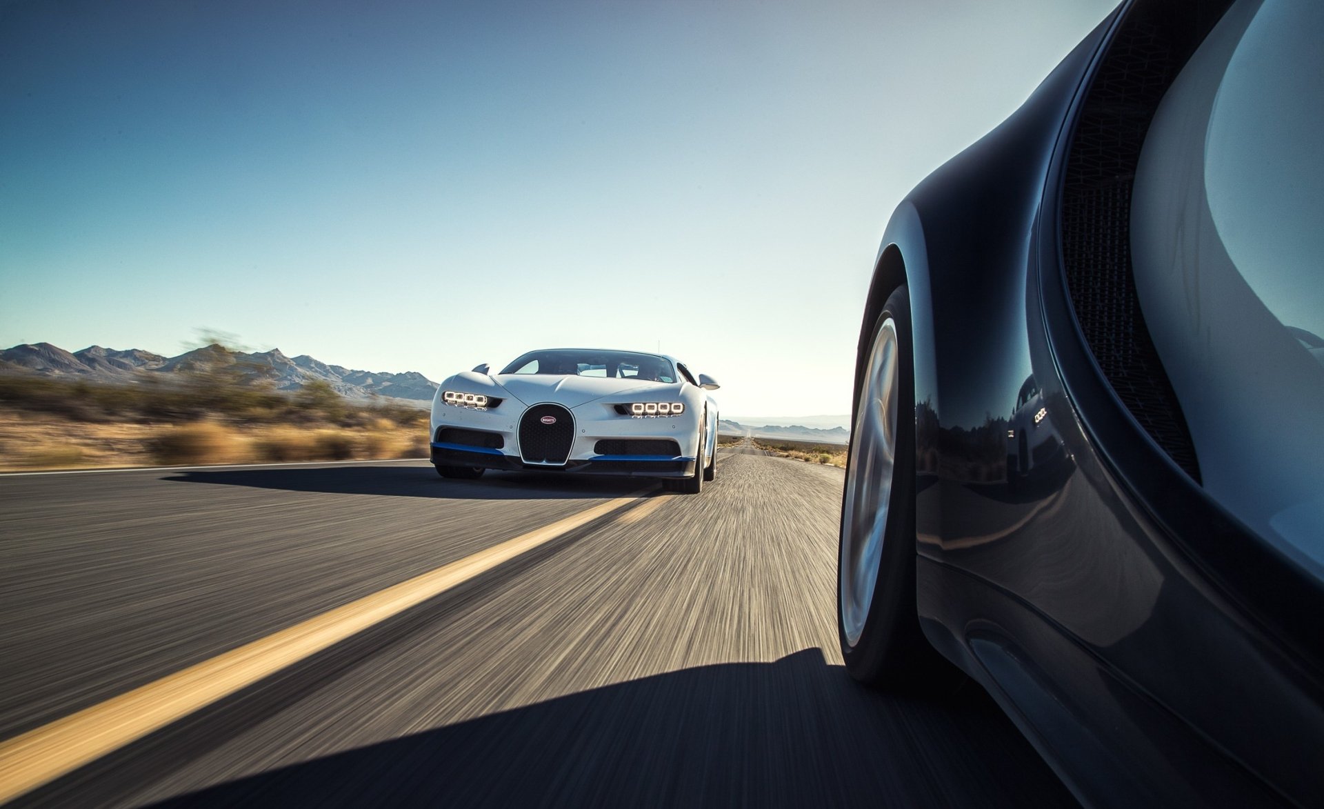 HD desktop wallpaper featuring a white Bugatti Chiron supercar speeding on an open road, captured from the perspective of a nearby vehicle.