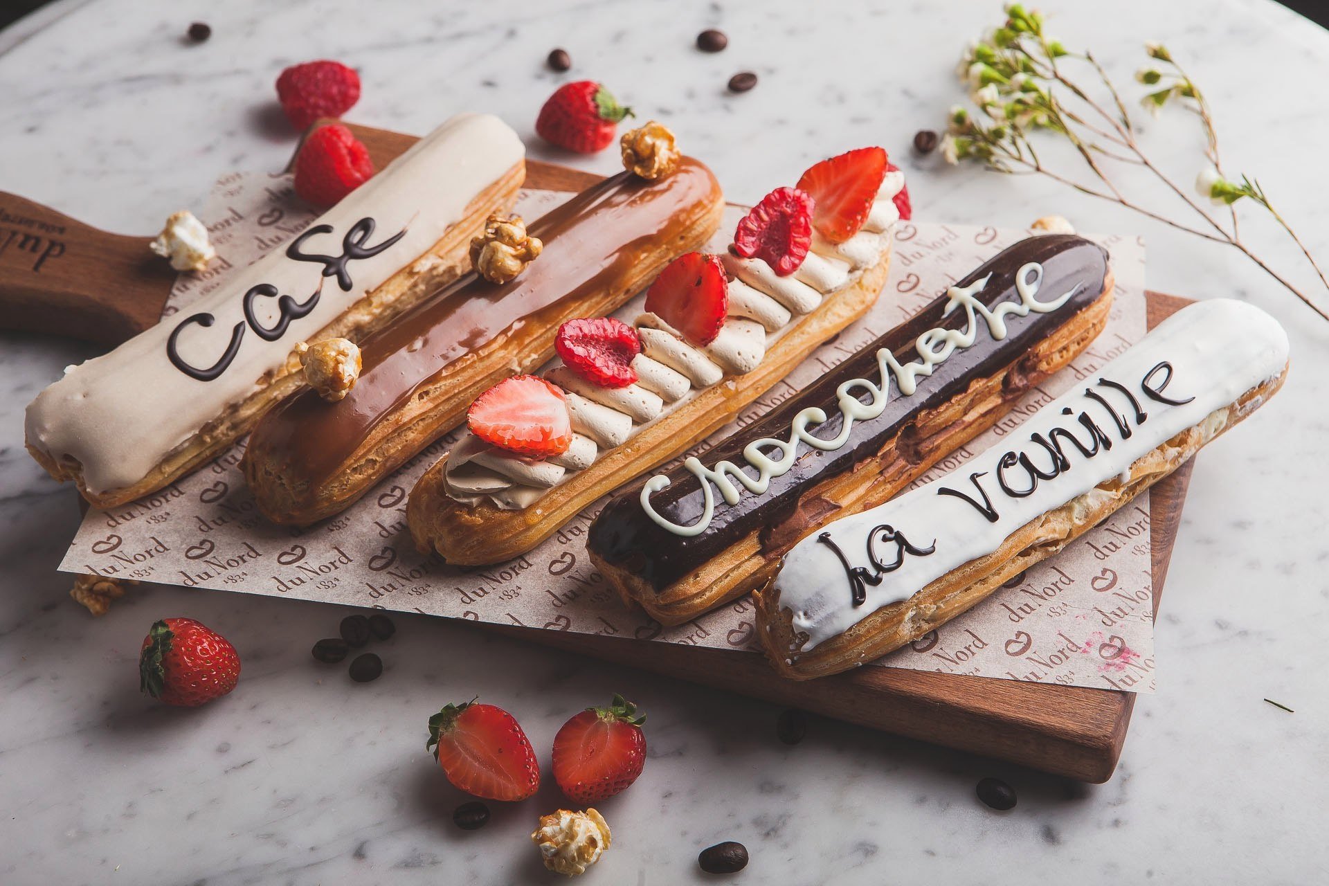 HD desktop wallpaper featuring a still life of four éclairs with café, chocolate, and vanilla flavors, adorned with fresh strawberries and nuts on a rustic wooden board.
